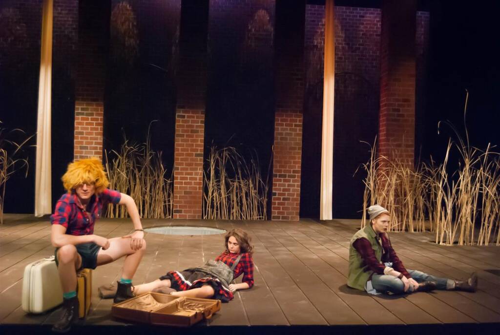 Three actors in plaid shirts perform on a stage with tall grasses and brick columns. One sits with a suitcase, another lies down, and the third sits cross-legged. The scene has a rustic, outdoor feel.