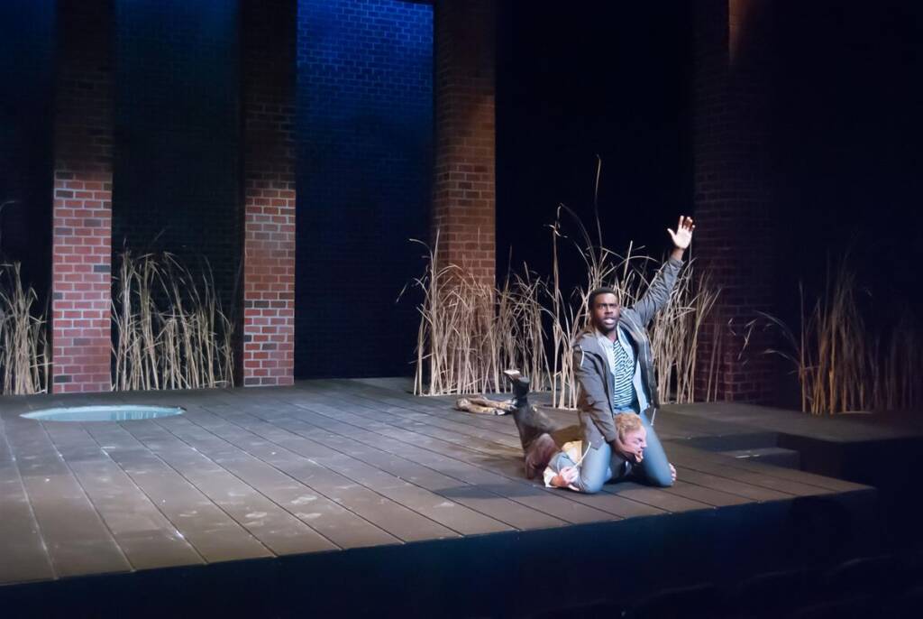 Two actors on a stage: one lies facedown, while the other, wearing a striped shirt and jacket, kneels beside him with an arm raised. The backdrop features tall plants and brick pillars, creating a dramatic atmosphere.
