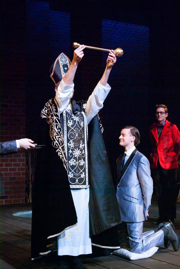 A person in ornate black and white ceremonial attire holds up a long object, facing a kneeling individual in a suit. Another person in a red jacket stands nearby, observing the scene. Beams of light illuminate the stage set against a dark background.