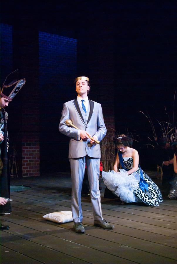 A person in a light gray suit stands confidently on a stage, holding a scepter and wearing a crown. Nearby, another person in a black and white gown sits or kneels. The scene appears to be part of a theatrical performance.