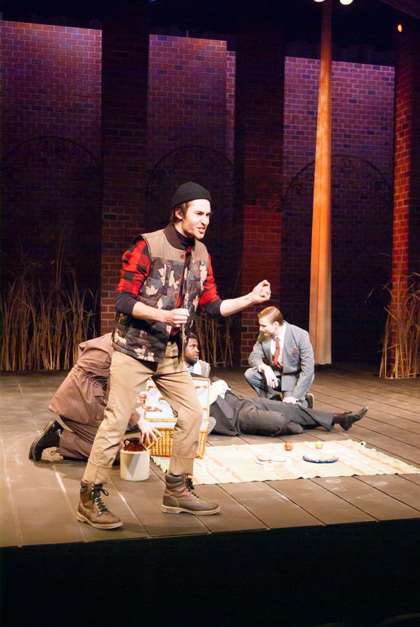 A stage performance features four actors. One stands energetically in the foreground wearing casual clothes and a beanie. Two actors sit, and one lies down on a picnic blanket with food props. The backdrop displays a brick wall with arches.