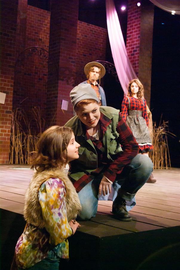 Four actors on a stage performing a play. Two are in the foreground; one is kneeling and smiling at the other, who is standing. In the background, two actors stand facing forward. The set includes brick walls and draped fabric.
