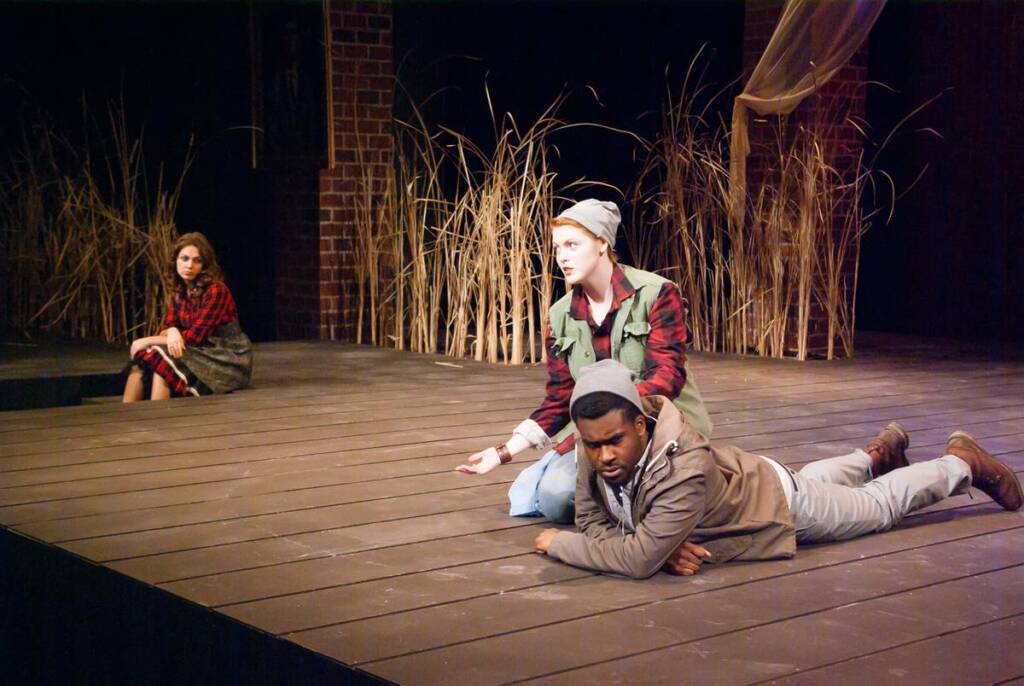Three actors perform on a stage with a rustic backdrop. Two sit close, one in a red and green jacket, and another lying down in a gray jacket. A third actor in a red plaid shirt sits apart. Tall reeds and brick walls are in the background.