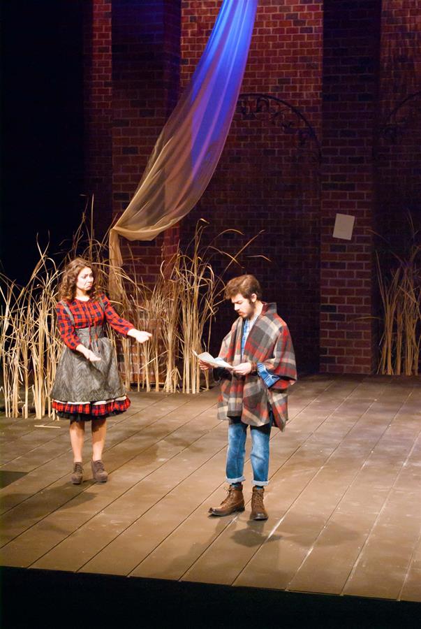 Two actors perform on a stage with a rustic backdrop and tall grass. The woman on the left wears a red plaid dress, while the man on the right wears a plaid poncho and jeans. A blue fabric drape hangs above under warm stage lighting.