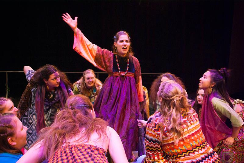 A woman in a colorful dress energetically gestures with one arm raised, surrounded by a group of people dressed in vibrant, patterned clothing. They appear engaged and attentive, with expressions of excitement.