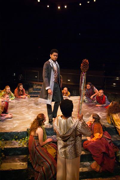 A theatrical performance with a man in period costume standing center stage, surrounded by seated actors in vibrant, eclectic attire. A person in the foreground raises a wand-like object. The stage is circular, with audience seating visible.