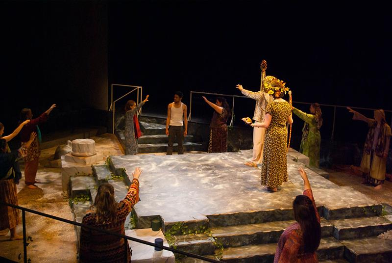 A theater scene with actors on a stone stage. One wears a flower crown and gestures outward, while others surround with raised arms. Their costumes suggest a mythological or ancient theme. The setting is dimly lit with a rustic, natural ambiance.