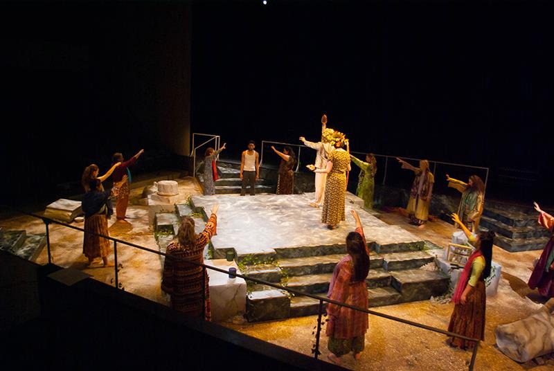 A theatrical performance on stage featuring actors in ancient-style costumes. They stand on a tiered stone structure, with a central figure holding a staff. The setting includes a sandy floor and dim lighting, creating an ancient or mythical atmosphere.