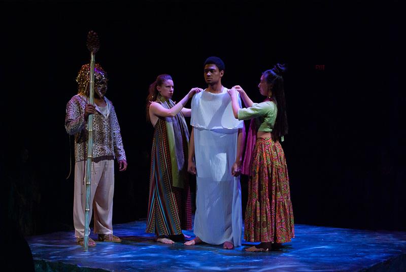 Four actors stand on a dimly lit stage. One wears a mask and holds a staff, while two others adjust a white robe on the fourth actor. The scene appears to be from a dramatic theater performance with cultural attire.