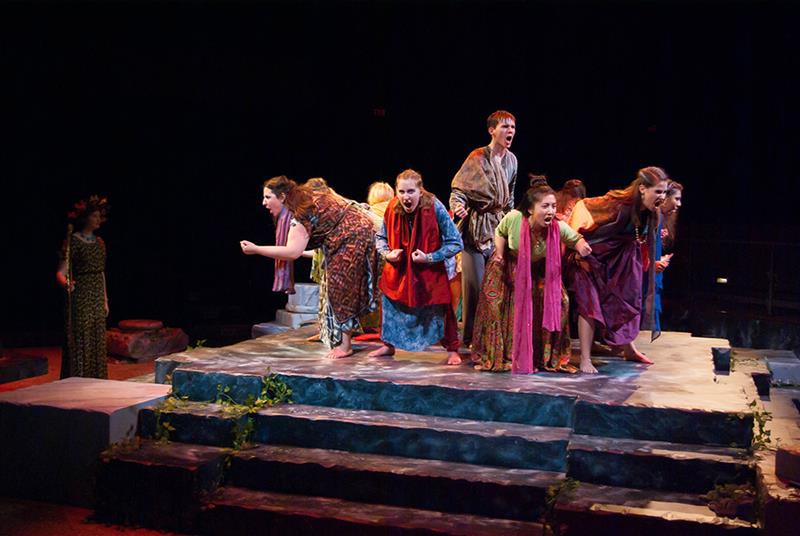 A group of actors wearing colorful costumes perform energetically on a stage with stone steps. They appear intense and animated, as if portraying a dramatic scene. The lighting creates a vivid, theatrical atmosphere.
