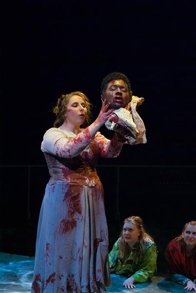 A woman in a blood-stained dress holds a prop head on stage, surrounded by other performers. The scene is dramatic and intense, set against a dark background.
