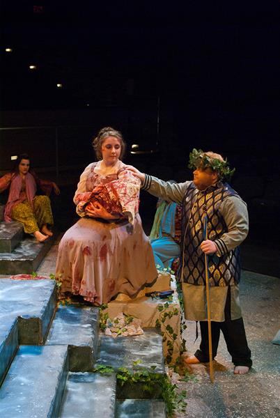 Two actors in theatrical costumes perform on stage. One is seated, dressed in a floral gown, holding a fan, with a contemplative expression. The other stands beside them, wearing a medieval-style outfit and a laurel crown, gesturing towards the seated actor.