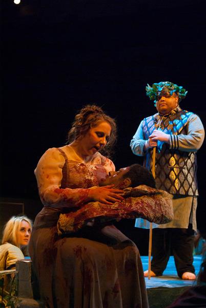 A woman in medieval attire holds a wounded man in her arms on stage. She has a solemn expression and the man appears lifeless with closed eyes. A person in a patterned robe and laurel crown stands behind them, holding a staff. Another woman watches from the side.