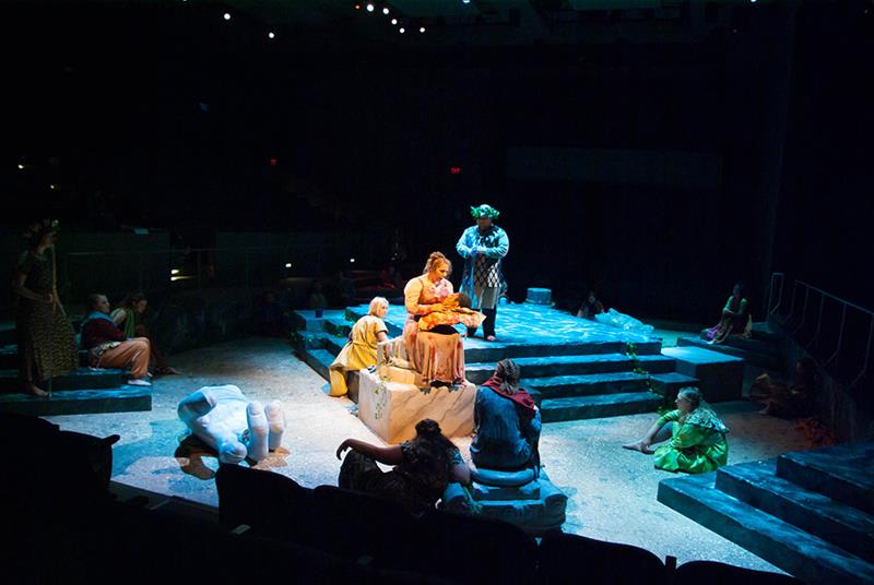 Stage play scene featuring actors in a dimly lit setting, dressed in period costumes. They are gathered on a stone-like platform with steps. One actor is seated holding a book, surrounded by others in various poses, with scattered props around.