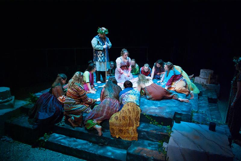 A group of people in colorful, patterned clothing are kneeling and sitting on a stone stage, focused on the center. One person stands slightly apart, wearing a crown of leaves. The scene is dimly lit, creating a dramatic atmosphere.