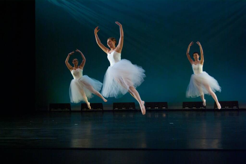 Three ballet dancers in white tutus leap gracefully on a dimly lit stage with a teal backdrop. Their arms are raised, and they appear in motion, creating a sense of elegance and harmony.