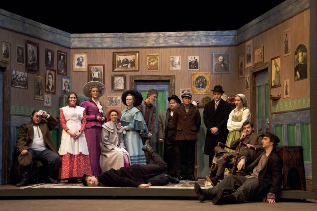 A group of twelve people in period costumes stand and sit in a room with vintage decor and framed photos. One person lies on the floor, while others pose around them. The room has painted walls and the scene resembles a theatrical play.