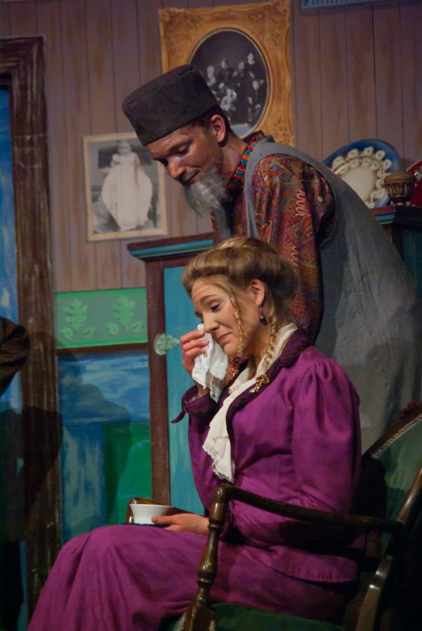 A woman in a purple dress sits on a chair, wiping her eyes with a handkerchief. A man in a hat and decorative clothing stands beside her with a comforting expression. They are in a room with framed photos and ornate decor.