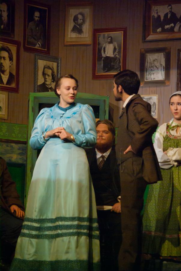 A theatrical scene with actors in period costumes. A woman in a blue dress stands beside a man in a brown suit. They are surrounded by a backdrop of framed portraits on a wooden wall, with another actor seated behind them.