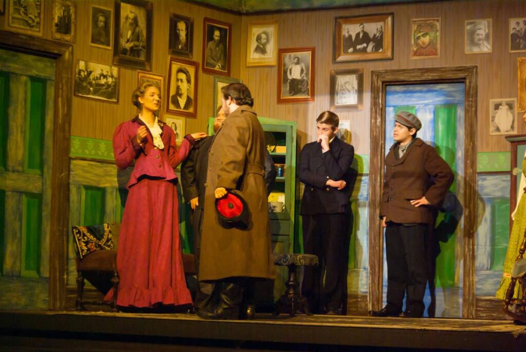 Five actors perform a theatrical scene on stage, dressed in period costumes. The set includes vintage framed photos on the walls, green doors, and a bookshelf. One actor gestures expressively, while the others listen attentively.
