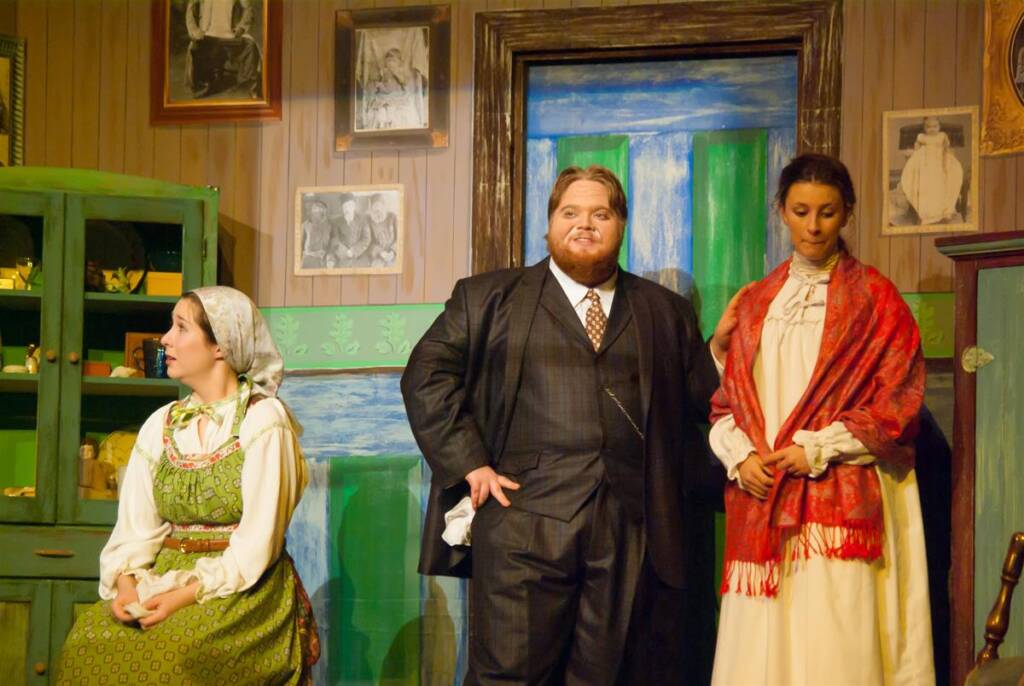 Three actors on stage dressed in period costumes. The man in the center is wearing a suit, while the women on either side are in traditional dresses. The backdrop includes family photos and painted scenery, suggesting a domestic setting.