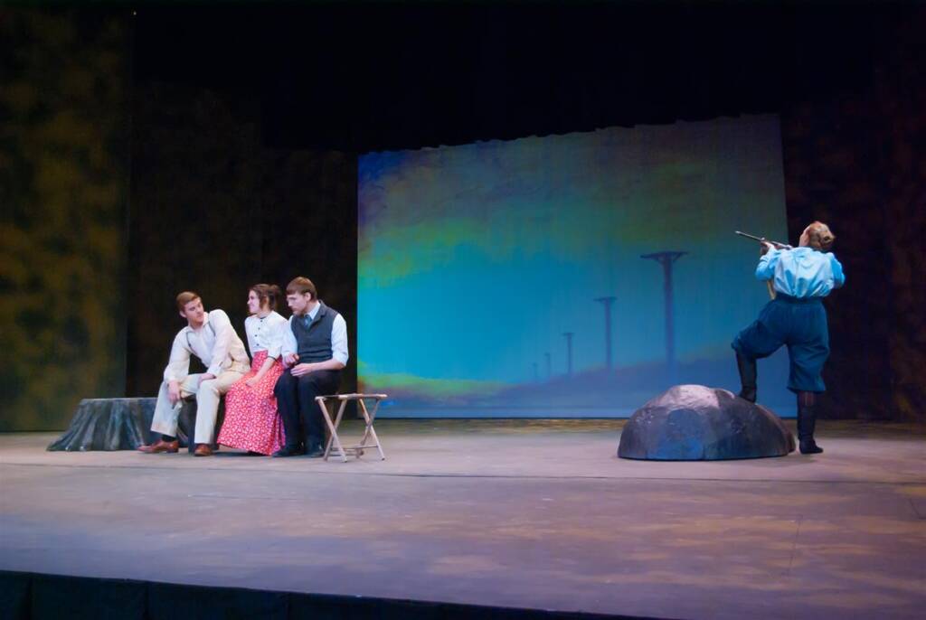 Four actors are on stage. Three are seated on the left, two men and a woman. One actor stands on the right, dramatically pointing a prop resembling a rifle. The background is a painted scene with vertical poles and a blue sky.