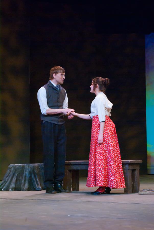 A man and a woman are performing on stage. The man, wearing a vest and trousers, holds hands with the woman, who is in a red floral skirt and white blouse. A bench and a tree stump prop are visible on the wooden stage floor.