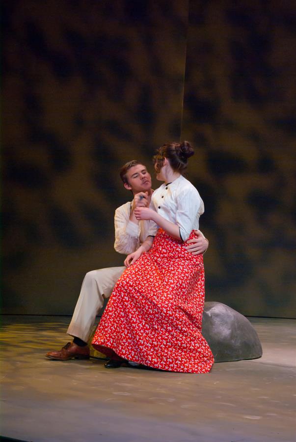 A man and woman are sitting on a rock on stage. The woman is wearing a long red skirt with a white blouse, and the man is dressed in light-colored pants and a white shirt. They are facing each other, with the man holding the woman's waist.