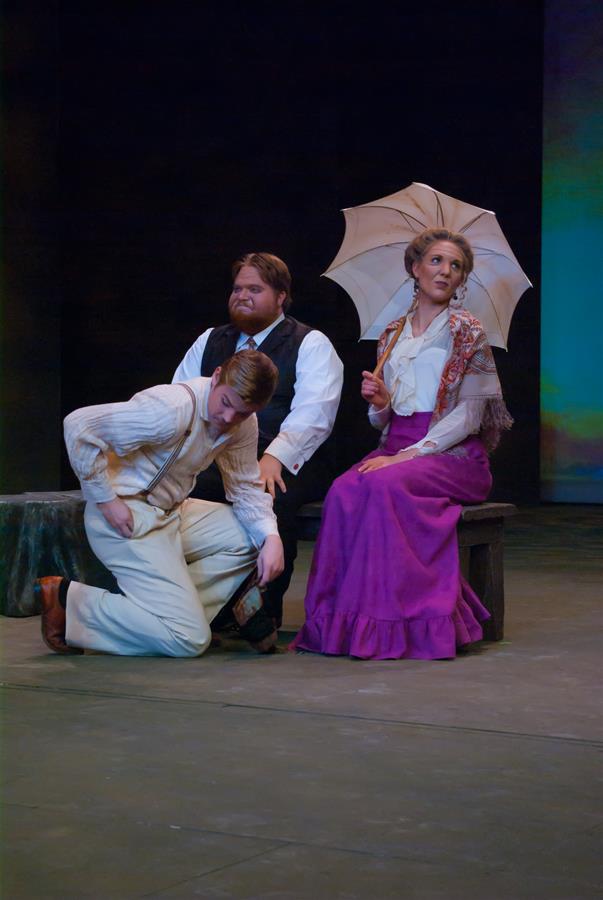 Three actors perform on stage. A man sits on a stump with a woman holding a parasol seated beside him; they both watch another man on one knee adjusting his shoe. The woman wears a purple skirt and shawl, the men wear vests and period attire.