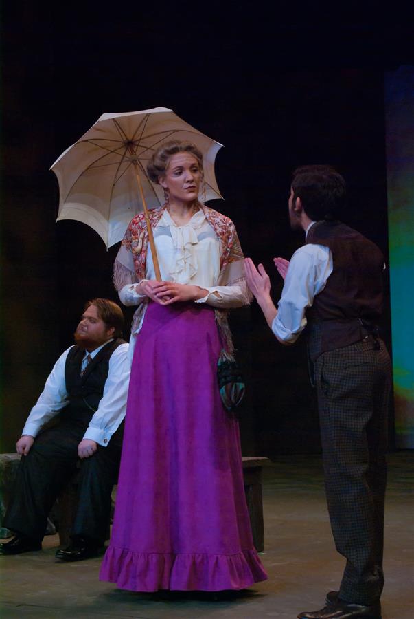 Three actors perform on stage. A woman in a long purple skirt and holding a parasol stands center, while a man in a vest gestures toward her. Another man sits on a bench in the background, listening. The setting suggests an early 20th-century scene.