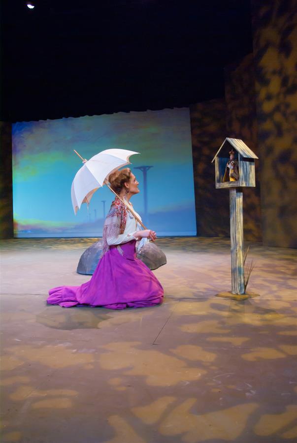 A woman in a purple dress kneels on a stage, holding a white parasol. In front of her is a small structure resembling a birdhouse on a stand. The background features a softly lit sky-like backdrop.