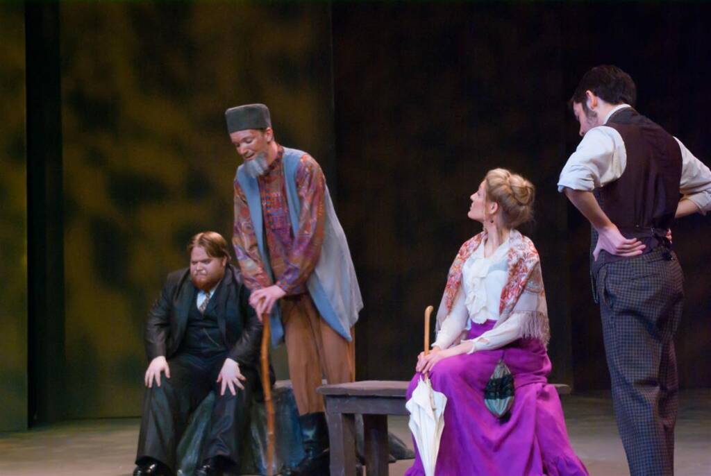 Four actors perform on stage in period costumes. A woman in a purple skirt sits on a bench, attentively listening to three men standing nearby. The scene is set against a dark, abstract background, suggesting a dramatic moment.