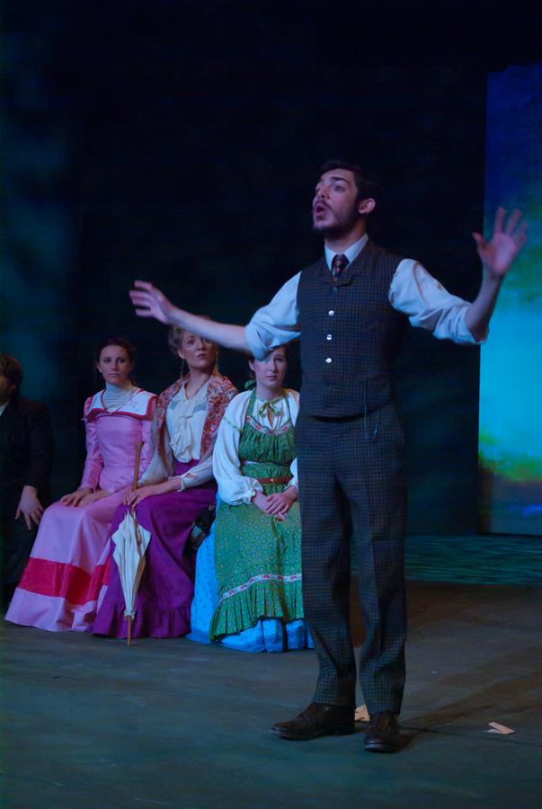 A person in a suit stands on stage with arms raised, passionately performing. Three others sit behind him in period costumes, watching attentively. The background is dark with a hint of a blue-lit scene.