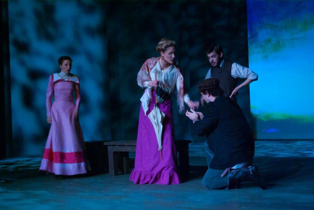 Four actors are performing on stage in period costumes. A woman in a bright magenta dress gestures emphatically, while a man kneels before her. Another woman and man watch from the side. The lighting casts dramatic shadows on the backdrop.
