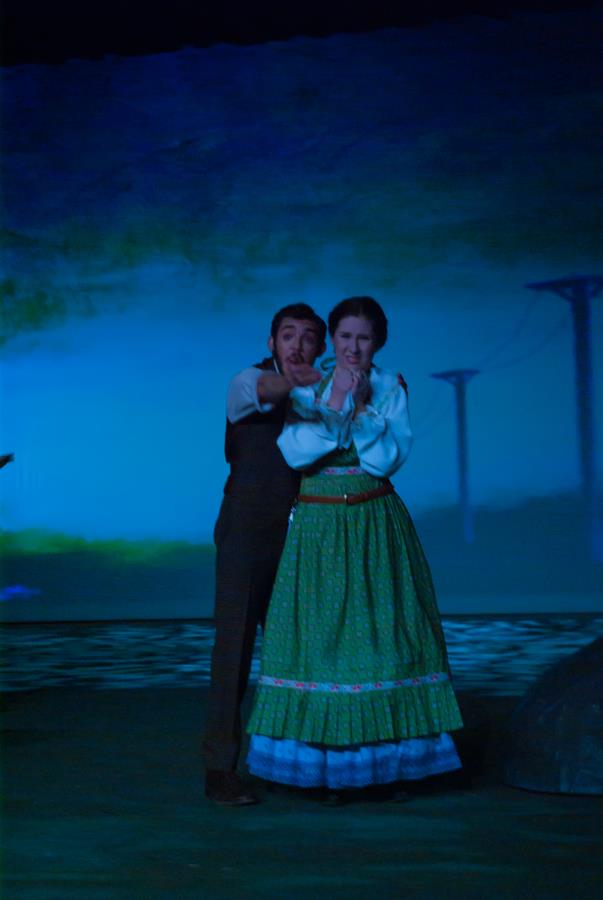 A man and woman in period costumes stand on a dimly lit stage, looking surprised or excited. The background is dark with subtle blue and green hues. The woman wears a green dress with a white blouse, while the man wears dark pants and a vest.
