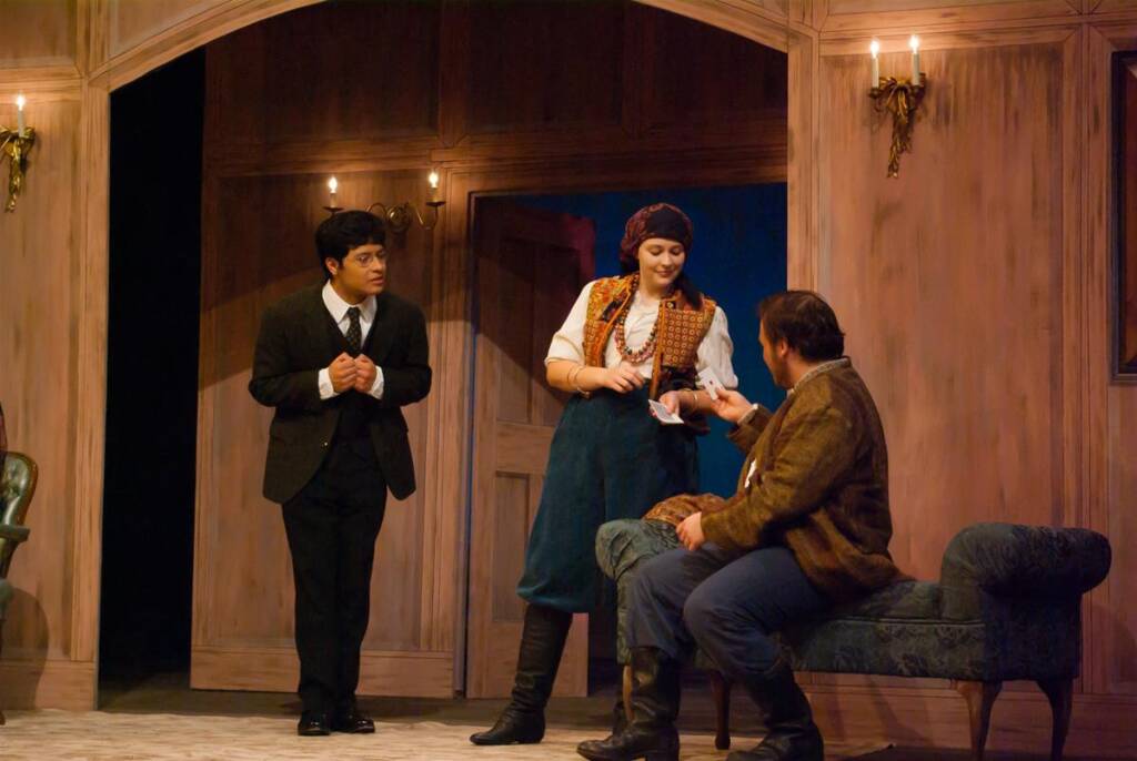 Three actors on stage in a theatrical setting. One man in a suit stands to the left, a woman in traditional attire with a headscarf stands center, and a seated man in rustic clothing gestures with a card. The backdrop is a warmly lit, wooden room.