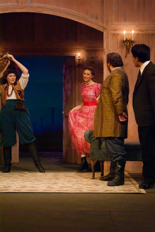 Four actors perform on stage in a period drama setting. One wears a rustic outfit, holding something overhead. Another in a pink dress stands by a door. Two men in suits look on. The backdrop suggests an evening scene.