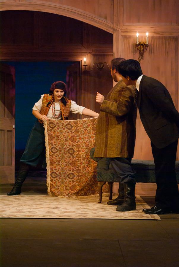 Three actors on stage in a theatrical scene: one person, wearing boots and a headscarf, holds a patterned rug. Two others, in formal attire, look towards the rug. They're in a room with wooden walls and soft lighting.