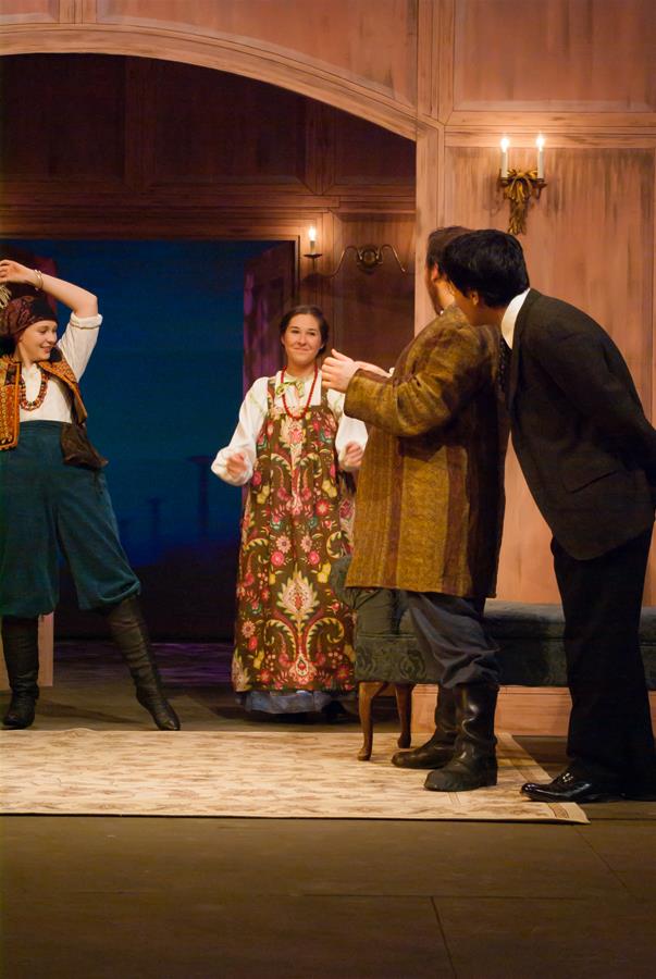 Four actors are performing on stage. One woman is wearing a colorful dress and gesturing, while the others, dressed in a variety of period costumes, are interacting with her. The setting features warm lighting and wooden paneling.