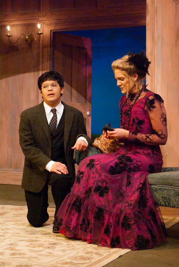 A man in a suit kneels on the floor, holding one hand to his chest, looking towards a seated woman. The woman is dressed in an elegant purple gown with black floral patterns, seated on a sofa, holding a small object. The setting is a room with wooden walls.