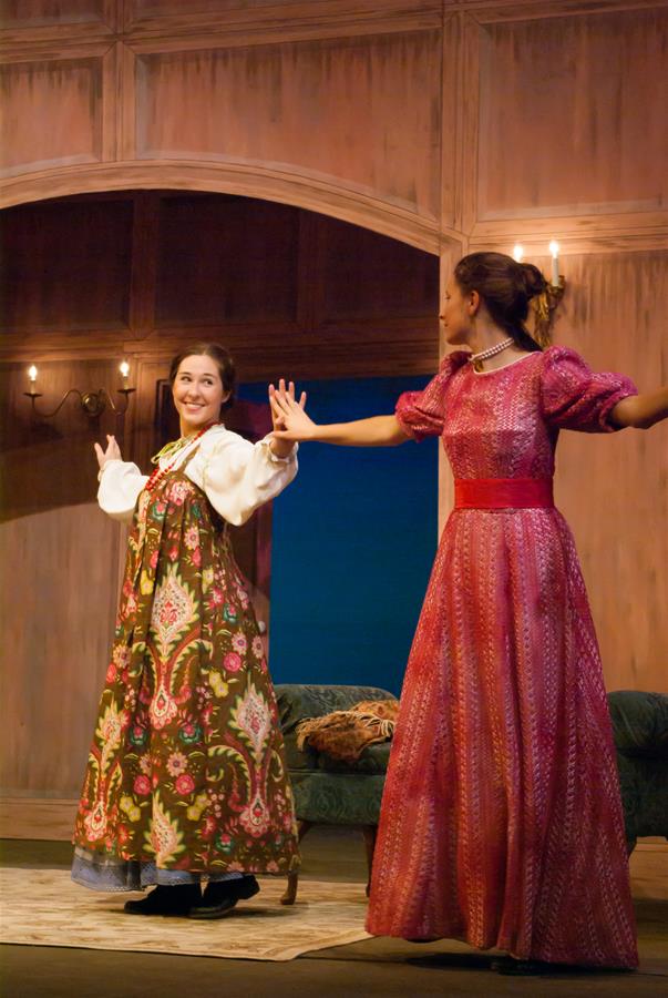 Two women are performing on stage in period costumes. One wears a floral-patterned dress, and the other is in a pink, patterned gown. They are holding hands and smiling, with a vintage-style room as their backdrop.