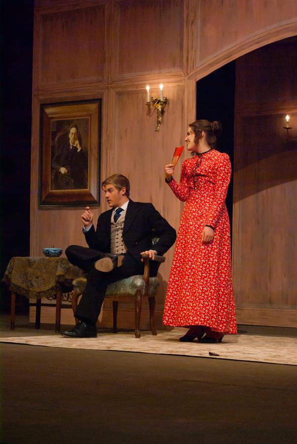 A man in a suit sits in a chair, looking up at a woman in a long red dress who is holding a book. They are on a stage set resembling a vintage living room, with a portrait on the wall and a table with a bowl.