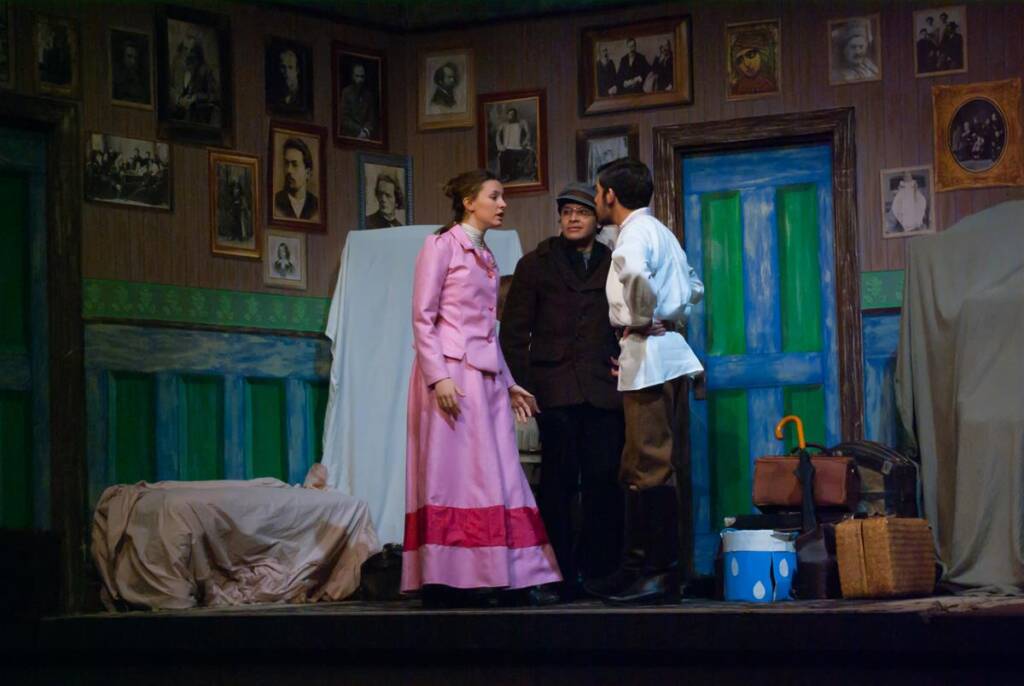Three actors perform on stage in a room with wooden walls, adorned with framed photos. One woman in a pink dress stands with two men wearing dark coats and boots. A bed with a draped sheet, colorful doors, and various props are in the background.