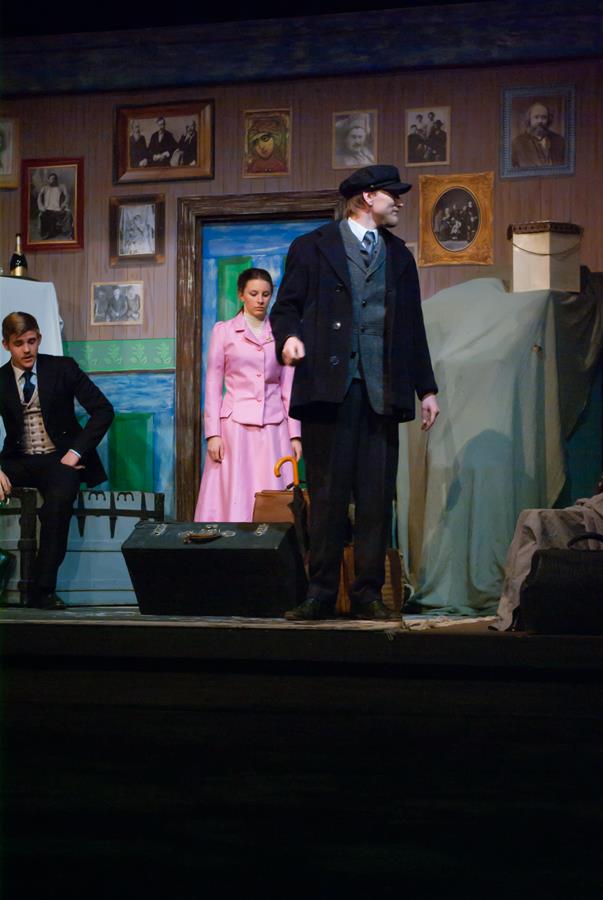 Stage scene with actors in period costumes. A man in a dark coat and cap stands prominently; a woman in a pink dress and a seated man in a suit are behind him. The set features vintage portraits and a covered object.