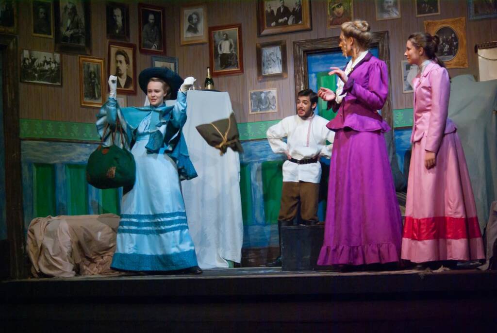 Four actors on stage in a theatrical performance. One in a blue dress holds a bag, while others in purple and pink dresses gesture dramatically. A man in a white shirt watches. The set features framed photos and a painted backdrop.