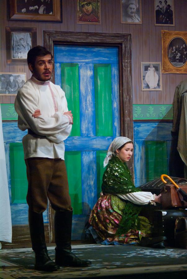 A theatrical scene with a man standing with arms crossed in front of a blue door. A woman in traditional clothing sits to his right, holding a suitcase and umbrella. The walls are decorated with old photographs.