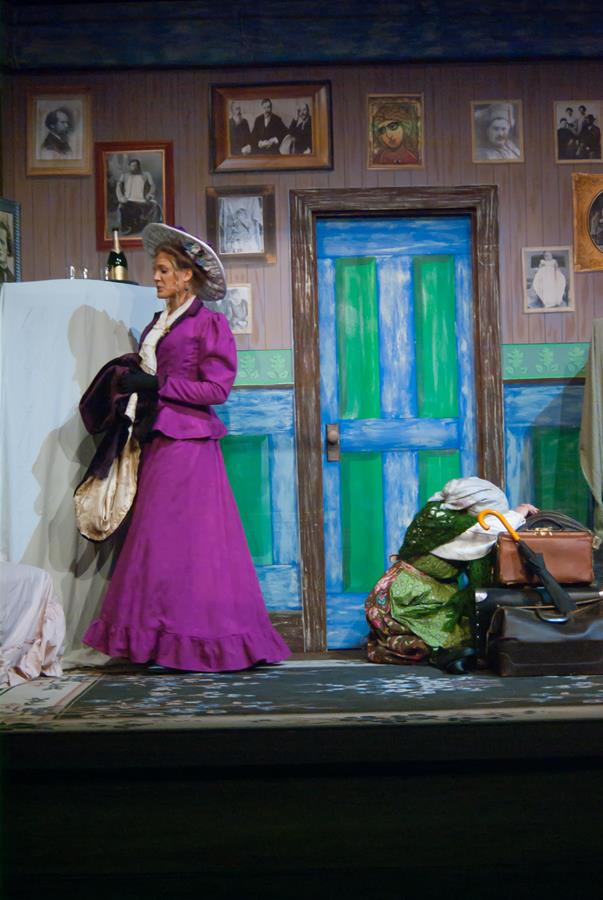 A woman in a purple Victorian-style dress and hat appears on stage. She stands near a bright blue and green door, holding some garments. Another person in a green dress is crouched by a suitcase. The stage is decorated with framed photos.