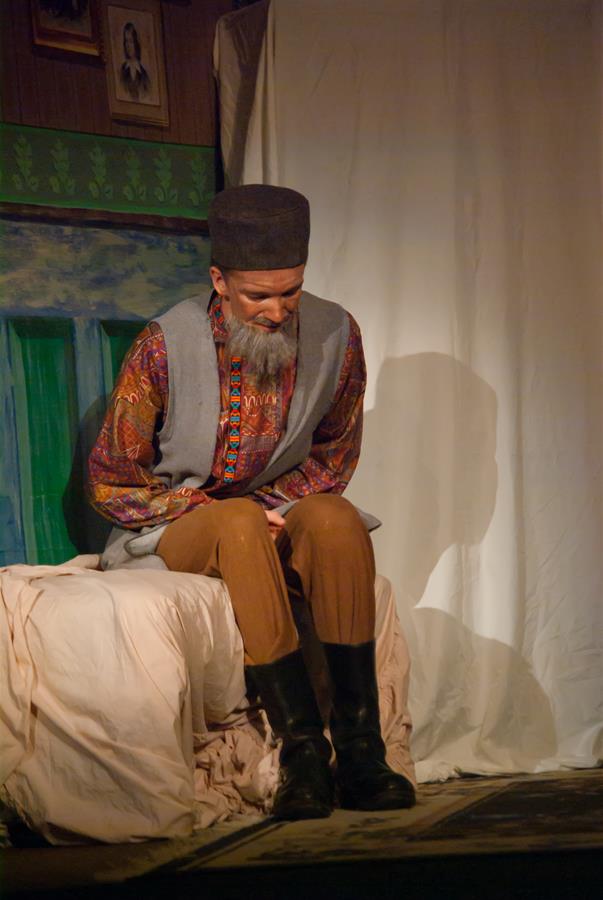 An older man with a gray beard sits on a bed, wearing a patterned shirt, vest, and brown pants with black boots. He wears a dark hat and looks downward, set against a backdrop featuring a framed picture and textured drapery.