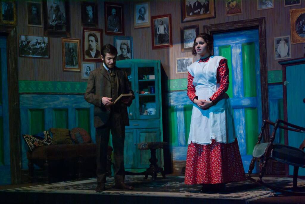 Two actors perform on stage. The room is decorated with numerous framed pictures on the walls. The man holds a book, wearing a suit, while the woman in a red dress and white apron stands nearby. The setting suggests a vintage, domestic interior.