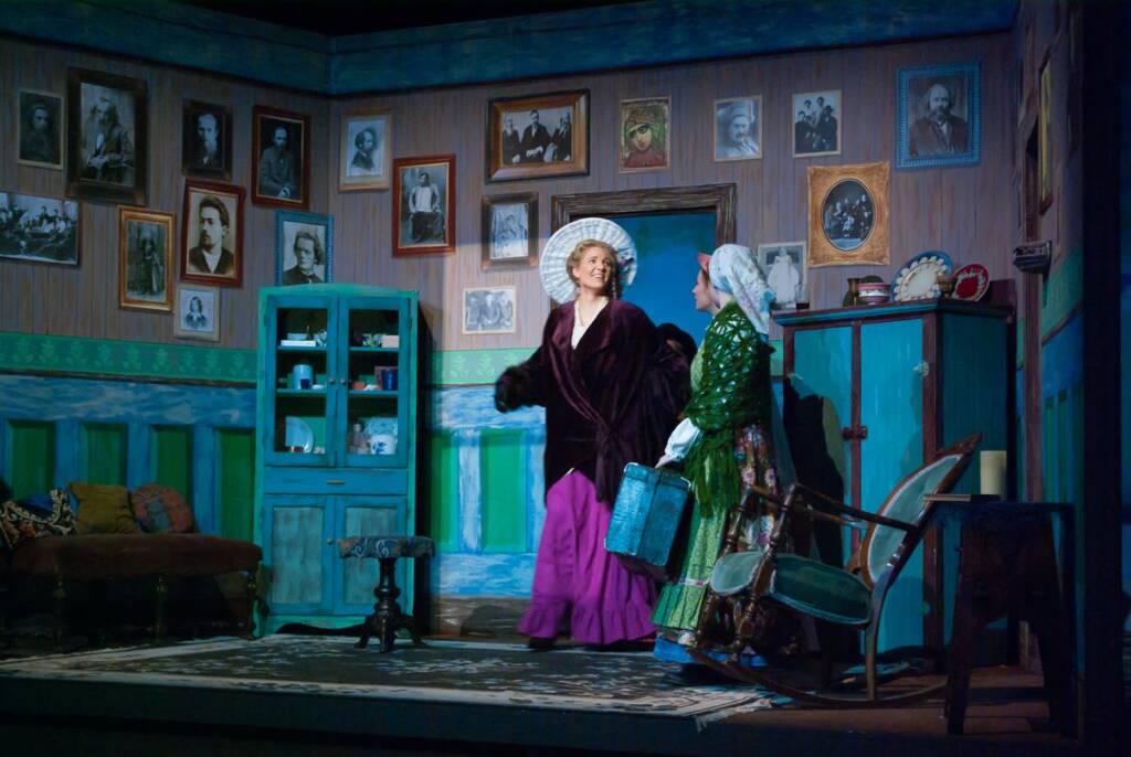 Two actors in period costumes on a stage set depicting a vintage living room. The room has wooden walls adorned with black-and-white photos, a blue cabinet, and various furnishings. One actor wears a bonnet and coat, while the other holds a basket.